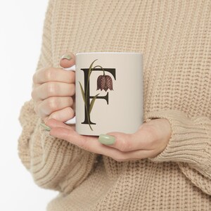 May include: A white ceramic mug featuring the letter "F" in a dark brown serif font, with a botanical illustration of a flower. The flower is a deep burgundy color with green leaves and stem. The mug is held in a person's hands.