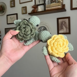 May include: Two handmade crochet turtles. One turtle has a sage green shell, the other has a yellow shell. Both turtles have light green heads and limbs, and black eyes. The turtles are held in front of a wall with framed art and a wooden shelf.