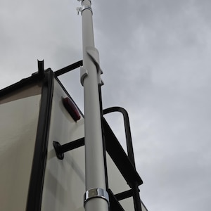 May include: A white aluminum telescoping flagpole mounted to the side of a recreational vehicle. The pole is secured with a black metal bracket and has a silver ring near the bottom.