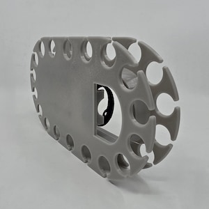 May include: A light grey, oval-shaped plastic organiser rack featuring a series of evenly spaced, U-shaped cutouts along its perimeter. The structure includes a central rectangular opening with a small black circular component visible inside.