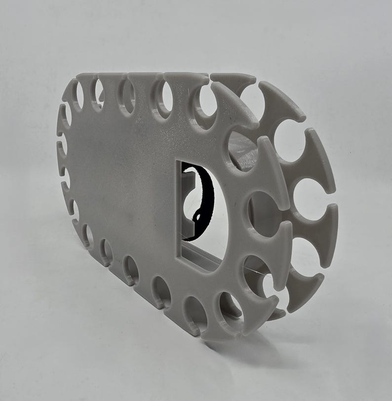 May include: A light grey, oval-shaped plastic organiser rack featuring a series of evenly spaced, U-shaped cutouts along its perimeter. The structure includes a central rectangular opening with a small black circular component visible inside.
