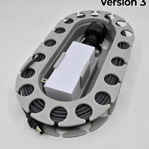 May include: An oval-shaped grey plastic cable organiser with a series of notches along the perimeter. A white rectangular power adapter with a black connector labelled 90w is secured in the centre, with a grey power cord coiled around the inner frame.