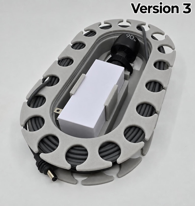 May include: An oval-shaped grey plastic cable organiser with a series of notches along the perimeter. A white rectangular power adapter with a black connector labelled 90w is secured in the centre, with a grey power cord coiled around the inner frame.