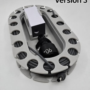May include: A grey oval cable organiser with a series of notches along the perimeter for winding a cord. The device holds a black power adapter labelled 90W and a white rectangular block. The text Version 3 is visible in the upper right corner.