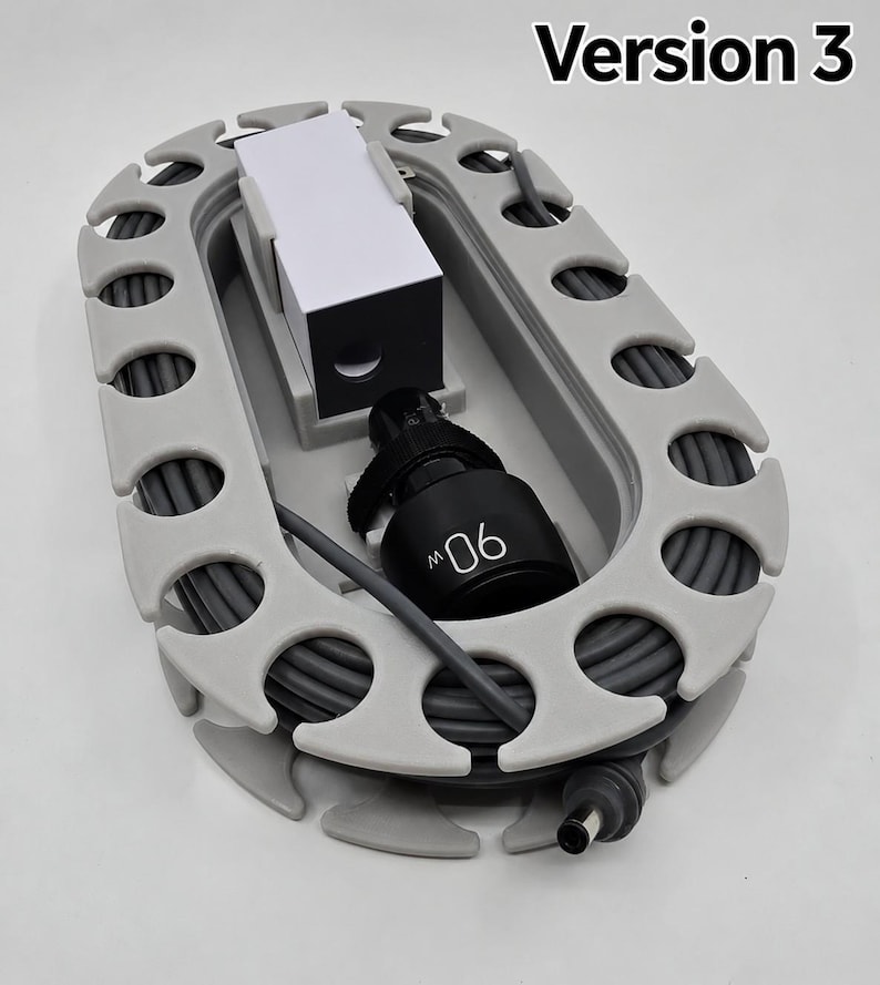 May include: A grey oval cable organiser with a series of notches along the perimeter for winding a cord. The device holds a black power adapter labelled 90W and a white rectangular block. The text Version 3 is visible in the upper right corner.