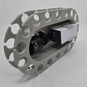 May include: A light grey oval storage rack with multiple circular cutouts along the perimeter. Inside the centre opening, a black cylindrical component labelled 90w is mounted next to a rectangular white and black block-shaped electronic device.