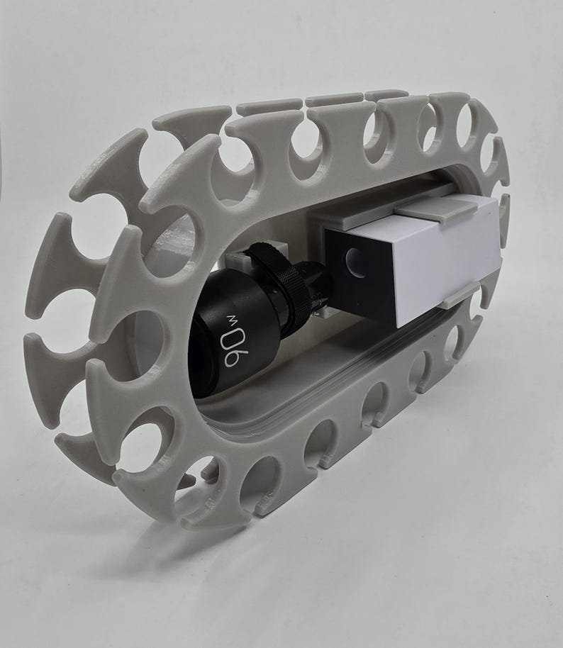 May include: A light grey oval storage rack with multiple circular cutouts along the perimeter. Inside the centre opening, a black cylindrical component labelled 90w is mounted next to a rectangular white and black block-shaped electronic device.