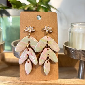 May include: A pair of dangle earrings with a floral design. The earrings are made of white and pink marbled clay with gold accents. The floral design is made of clear rhinestones.