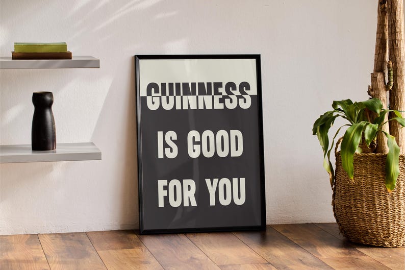 May include: A framed black and white print with the text "GUINNESS IS GOOD FOR YOU" in a bold, sans-serif font. The print is displayed on a wooden floor, with a plant in a woven basket and a decorative shelf in the background.