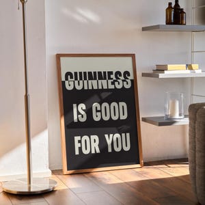May include: A framed print with the words "Guinness is Good For You" in white text on a black background. The frame is a light brown color. The print is leaning against a white wall.