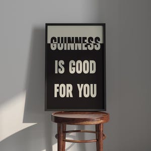 May include: A framed print with the text "GUINNESS IS GOOD FOR YOU" in white letters on a black background. The frame is black, and the print is displayed on a wooden stool.