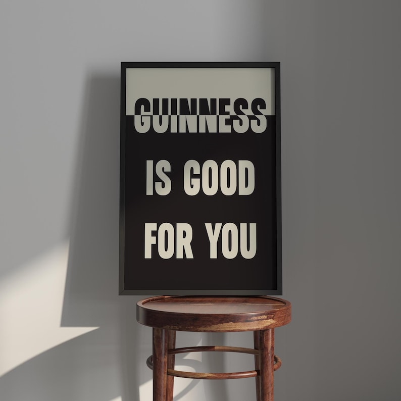 May include: A framed print with the text "GUINNESS IS GOOD FOR YOU" in white letters on a black background. The frame is black, and the print is displayed on a wooden stool.