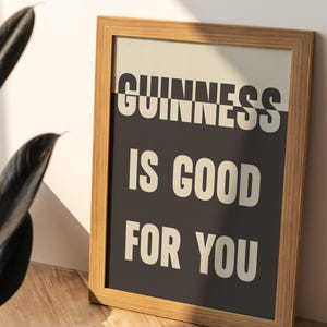 May include: Wooden framed print with the text "GUINNESS IS GOOD FOR YOU" in white letters on a black background. The print is leaning against a wall, with a plant in the foreground.