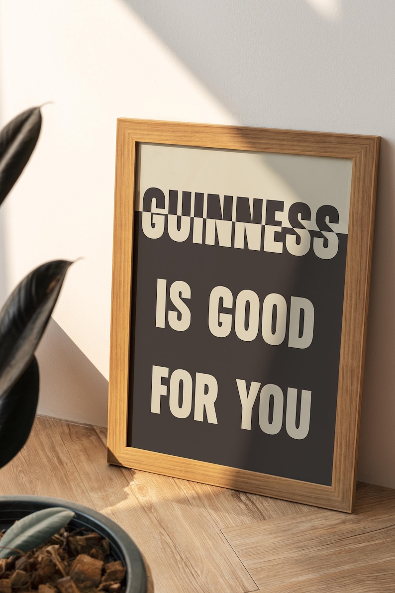 May include: Wooden framed print with the text "GUINNESS IS GOOD FOR YOU" in white letters on a black background. The print is leaning against a wall, with a plant in the foreground.