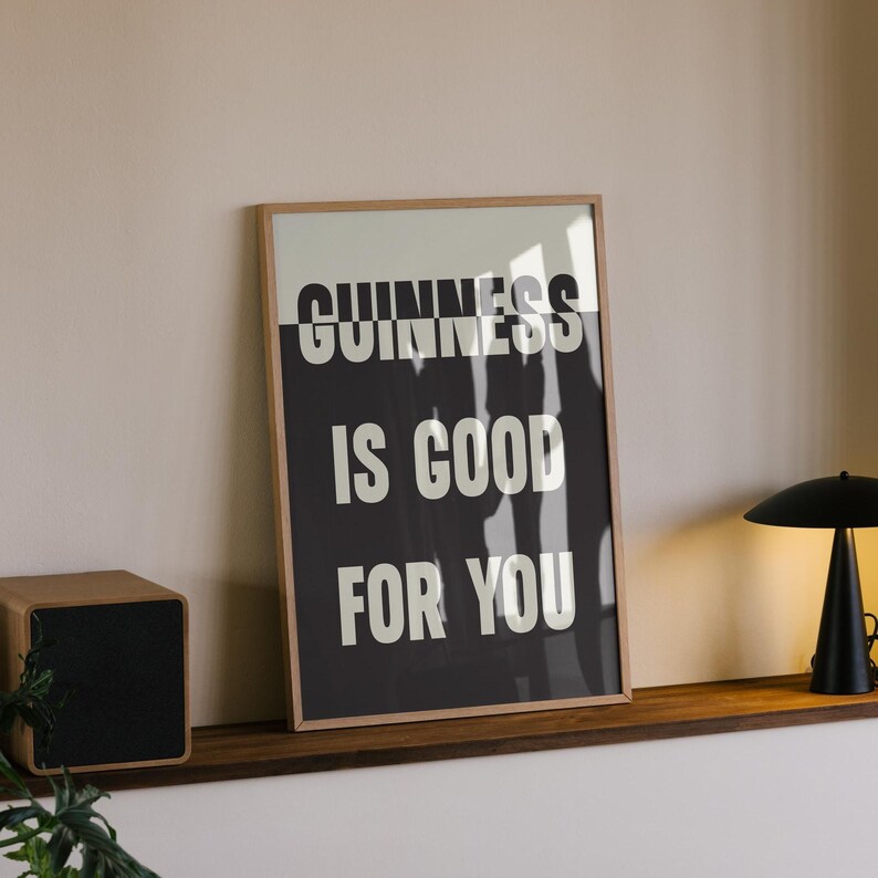 May include: Framed print with the text "GUINNESS IS GOOD FOR YOU" in white letters on a black background. The print is in a wooden frame and is displayed on a wooden shelf. A small black lamp and a wooden box are also on the shelf.