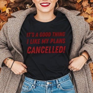 May include: Black t-shirt with the text "IT'S A GOOD THING I LIKE MY PLANS CANCELLED!" in red glitter lettering. The person is wearing blue jeans and a gray coat, lying on a bed of autumn leaves.