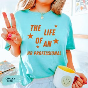 May include: A light blue t-shirt with the text "THE LIFE OF AN HR PROFESSIONAL" in orange glitter lettering. The shirt has a crew neck and short sleeves. A white mug with a yellow interior and a smiley face is visible.