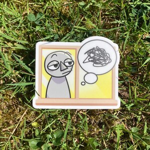Pu&ograve; includere: Un adesivo raffigurante un personaggio dei cartoni animati che guarda fuori da una finestra gialla e marrone, con una nuvoletta che mostra un disegno scarabocchiato. Il personaggio &egrave; grigio. L'adesivo &egrave; su una superficie erbosa.