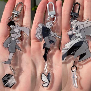 May include: Three clear acrylic keychains featuring anime-style characters in shades of gray. Each keychain has a silver-tone clasp. One keychain has a geometric shape, another a butterfly, and the last a sword.