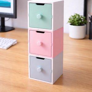 Può includere: Una pila di tre piccoli cassetti quadrati portaoggetti dai colori pastello. Il cassetto superiore è verde menta, quello centrale rosa e quello inferiore grigio. Ogni cassetto ha un piccolo pomello rotondo e una maniglia incassata. I cassetti sono impilati su una superficie di legno.