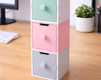 May include: A stack of three small, square storage drawers in pastel colors. The top drawer is mint green, the middle is pink, and the bottom is gray. Each drawer has a small, round knob and a recessed handle. The drawers are stacked on a wooden surface.