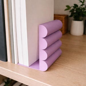 Könnte beinhalten: Ein lilafarbener Buchstütze hält einen Stapel Bücher auf einem Holzregal. Die Buchstütze hat ein geschichtetes, gebogenes Design. Die Bücher sind in verschiedenen Farben. Das Regal ist aus hellem Holz.