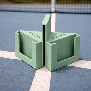 Può includere: Un'attrezzatura sportiva triangolare verde chiaro con la scritta "Vemme" stampata sopra. Il pezzo è realizzato in un materiale solido e ha una fessura al centro. È appoggiato su una superficie blu con linee bianche.