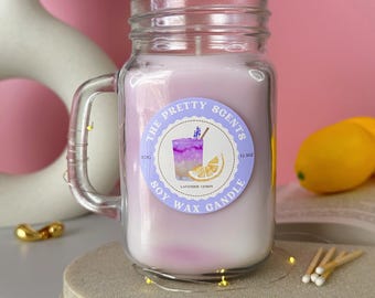 May include: A lavender-scented soy wax candle in a mason jar with a handle. The candle has a label that reads "The Pretty Scents" and "Lavender Lemon." The jar is on a gray platform with fairy lights, with lemons and a vase of lavender in the background.