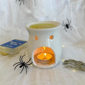 May include: A white ceramic wax warmer with a lit tea light candle inside. The warmer has two small holes and a larger opening. Black spider decorations and a skeleton hand are placed around the warmer, suggesting a Halloween theme.