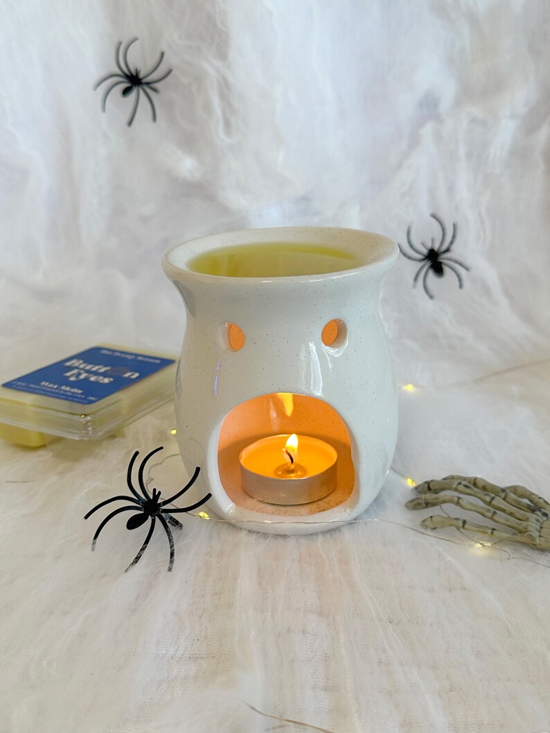 May include: A white ceramic wax warmer with a lit tea light candle inside. The warmer has two small holes and a larger opening. Black spider decorations and a skeleton hand are placed around the warmer, suggesting a Halloween theme.