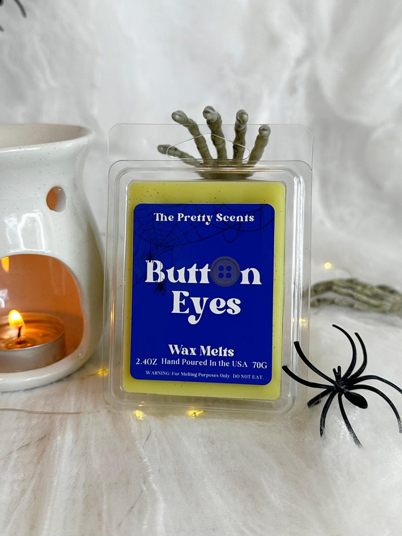 May include: A yellow wax melt package with the text "Button Eyes" and a spider web design. The package includes a plastic container with a skeleton hand and a black plastic spider. The wax melt weighs 2.4 oz (70g).