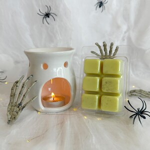May include: A white ceramic wax warmer with a lit tealight candle inside, next to a package of six green wax melts. Decorative skeleton hand and spider accents are placed on a white web-like background, creating a Halloween theme.