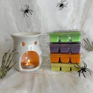 May include: A white ceramic wax warmer with a lit tealight candle, next to stacks of colorful wax melts in green, purple, orange, and yellow. The scene is decorated with spiderwebs, spiders, and a skeleton hand, creating a Halloween theme.
