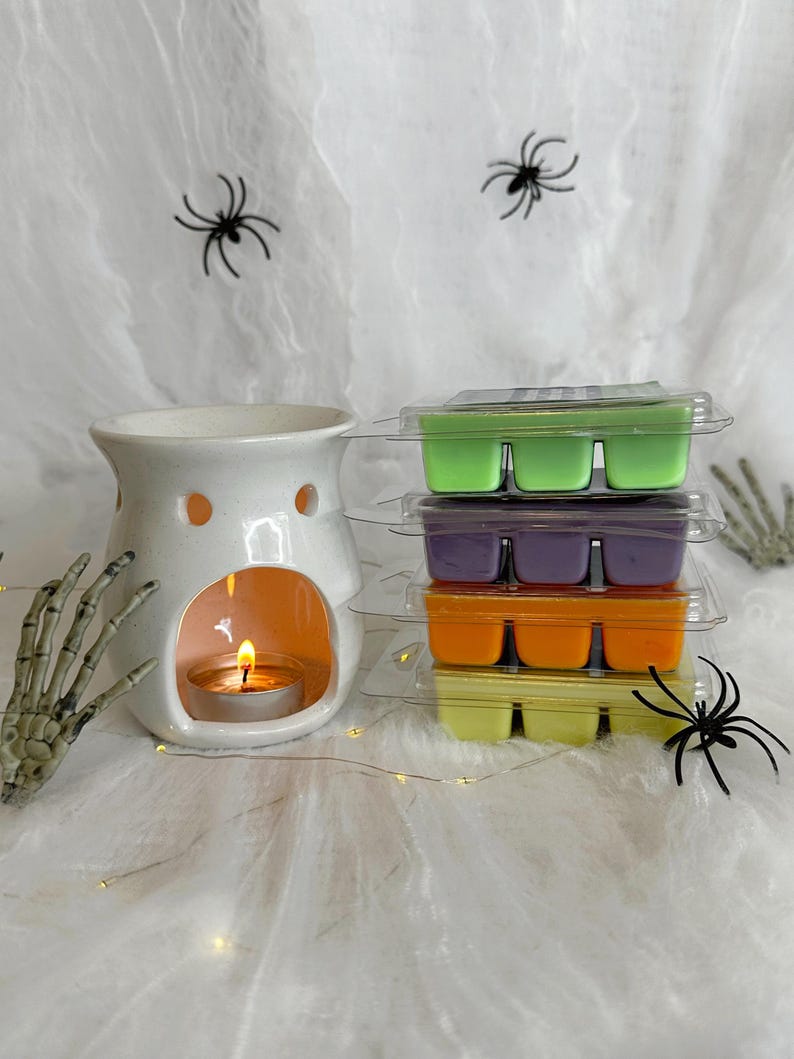 May include: A white ceramic wax warmer with a lit tealight candle, next to stacks of colorful wax melts in green, purple, orange, and yellow. The scene is decorated with spiderwebs, spiders, and a skeleton hand, creating a Halloween theme.