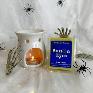 May include: A Halloween-themed image featuring a white ceramic wax warmer with a lit candle, a package of "Button Eyes" wax melts, and decorative spiders and a skeleton hand. The wax melts are in a clear plastic container with a blue label.