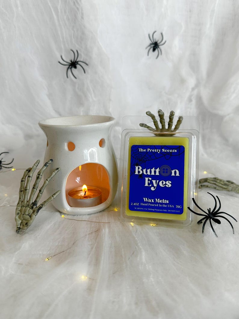 May include: A Halloween-themed image featuring a white ceramic wax warmer with a lit candle, a package of "Button Eyes" wax melts, and decorative spiders and a skeleton hand. The wax melts are in a clear plastic container with a blue label.