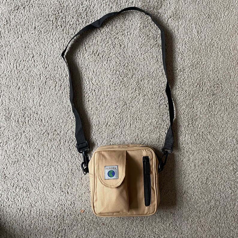 May include: A tan canvas crossbody bag with a black adjustable strap. The bag has a zipped pocket on the front and a small patch with the word "Pangea" and a globe logo.