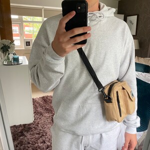May include: A person wearing a light grey hoodie with a black drawstring and a beige crossbody bag with a black strap.