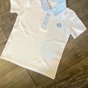 May include: White polo shirt with a blue gingham collar and a three-letter monogram in light blue. The shirt has short sleeves and a buttoned placket. The shirt is made of a soft, comfortable material.