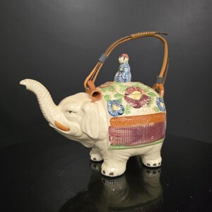 Peut inclure: Théière en céramique crème en forme d'éléphant, avec une anse tressée et une petite figurine sur le dos. L'éléphant est décoré de motifs floraux bleus, rouges et verts. La trompe est levée et le bec est orange.