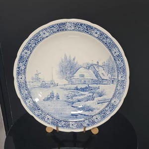 Puede incluir: Plato decorativo de cerámica blanca con una escena de paisaje azul y blanco. El plato tiene un borde festoneado con un diseño floral y de desplazamiento azul. El paisaje representa un molino de viento, casas y figuras cerca del agua.
