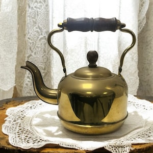 May include: A vintage brass kettle with a wooden handle. The kettle is sitting on a white lace doily on a wooden surface.