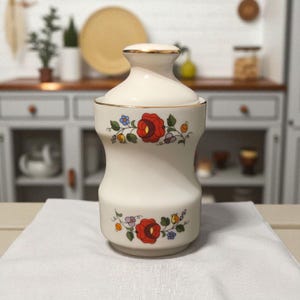 May include: A cream-coloured ceramic jar with a lid, featuring a gold rim and floral patterns. The jar displays a red rose and colourful flowers. The jar is likely for storage and is placed on a white cloth.