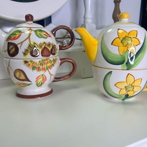 Op de afbeelding: Twee keramische theepotten met bijpassende theekopjes. De theepot links heeft een bruin handvat en een herfstbladerenontwerp met bruine, oranje en gele bladeren. De theepot rechts heeft een geel handvat en een narcissenontwerp met gele bloemen en groene bladeren.