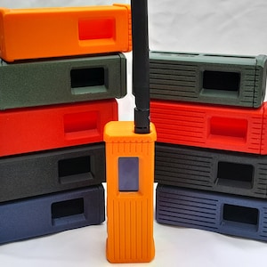 May include: A stack of eight colourful radio cases in various shades of green, orange, red, black, and blue. The cases are made of a textured material and have a rectangular shape. A single orange radio with a black antenna is in front of the stack.