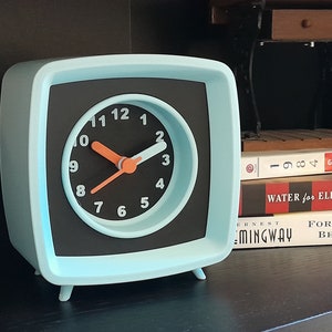 Puede incluir: Un despertador retro azul claro con agujas negras y detalles naranjas. La esfera del reloj tiene números negros y fondo blanco. El reloj está sobre una estantería con libros.
