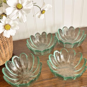 May include: Four clear glass bowls with a flower petal design are arranged on a wooden surface. The bowls have a light green tint. A wicker basket with white flowers is in the background.