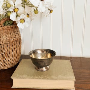 May include: A small, polished silver-toned metal bowl with a rounded body and a circular pedestal base. The bowl features a smooth, reflective surface and sits centered on top of a thick, light olive-green hardcover book.