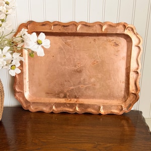 May include: A rectangular copper tray featuring a scalloped edge and a smooth, metallic surface with visible natural patina and light surface marks. The tray has a raised border and a flat base, showcasing the warm, reddish-brown tone of the metal.