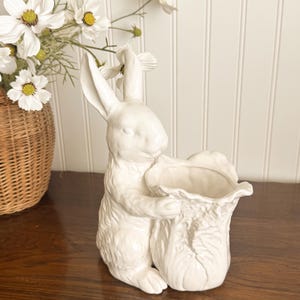 May include: A white ceramic bunny figurine holding a textured vase. The bunny has long ears and is hugging the vase, which has a ruffled top. The figurine is on a wooden surface, with white flowers in a wicker basket in the background.